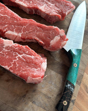 Tanzo Cutlery Kaizen utility knife with teal dyed wood and black resin handle with gold star mosaic pins resting beside raw marbled beef short ribs on a wooden cutting board