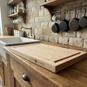 Tanzo Cutlery cherry wood cutting board with dual juice grooves and laser engraved logo resting on a rustic dark wood kitchen counter beside a cast iron skillet on a gas stove and a spice rack filled with seasonings