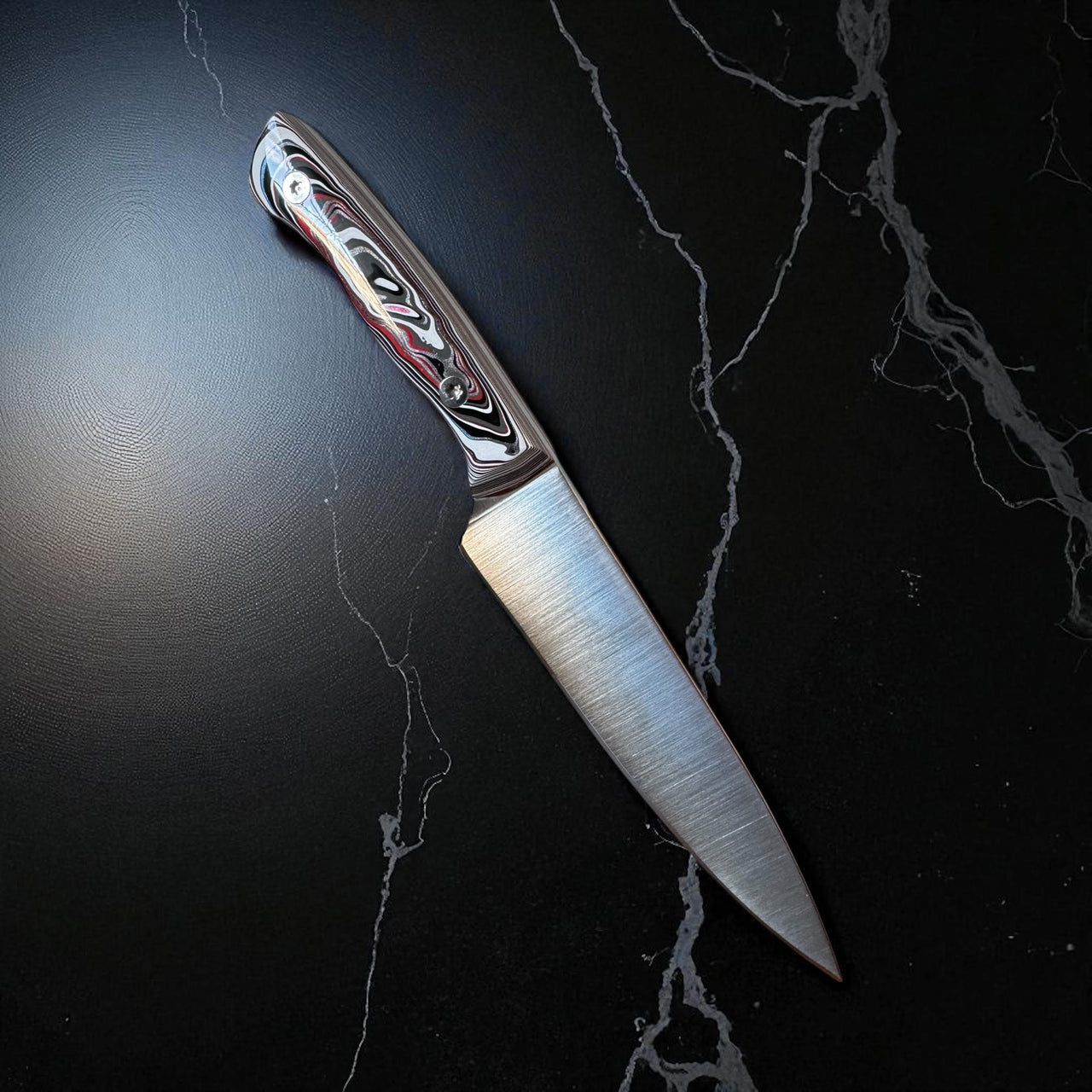  Tanzo Cutlery Kaizen 6 inch handcrafted utility knife with black white and red Fordite handle and brushed AEB-L stainless steel blade resting diagonally on a black marble surface