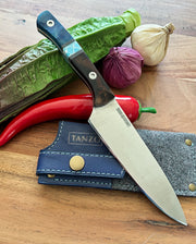 Tanzo Cutlery Kaizen utility knife with dark burl wood and blue turquoise resin handle with star pattern stainless steel screws resting on a navy blue TANZO leather sheath and gray felt liner on a wooden surface with bitter melon, red chili pepper, red onion, and white onion