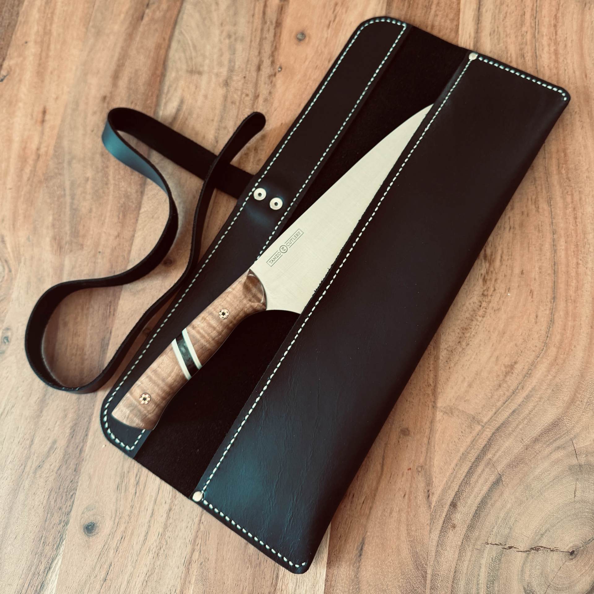 TANZO CUTLERY professional chef knife with wooden handle and black leather sheath on a wooden surface